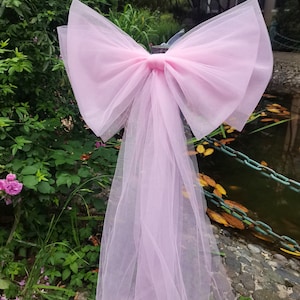 Puede incluir: Un gran lazo decorativo de color rosa claro confeccionado en tela de tul transparente. El lazo presenta dos bucles anchos y voluminosos junto con dos largas cintas que caen hacia abajo, creando un aspecto suave y superpuesto con una textura translúcida.