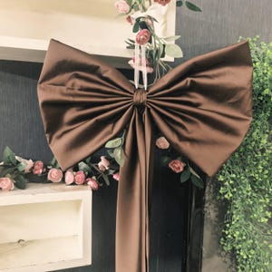 May include: A large, dark brown satin bow featuring a smooth, reflective fabric surface with prominent pleats radiating from the centre knot. Two long, straight fabric tails extend downwards from the central knot of the bow.