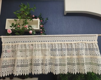 May include: A white lace valance with a scalloped bottom edge. The valance has a delicate pattern of diamonds and flowers. It is hung on a white rod.