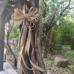 Puede incluir: Un gran lazo marrón hecho de cinta de arpillera está atado alrededor de una gruesa vid que crece en un poste de madera. La vid está envuelta varias veces alrededor del poste y está asegurada con cuerda. El lazo está atado en un nudo grande y suelto y los extremos de la cinta se dejan largos y flotantes. El lazo es un elemento decorativo que añade un toque de encanto rústico al poste.