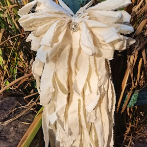 Puede incluir: Un lazo de tela blanco con un gran strass dorado brillante en el centro. El lazo está atado alrededor de un poste verde y cuelga en largas tiras deshilachadas.