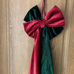 May include: A large, velvet bow in Christmas colors of red and green hangs against a wooden door. The bow is made of two colors of velvet, with a red center and green accents. A red ribbon is used for hanging.