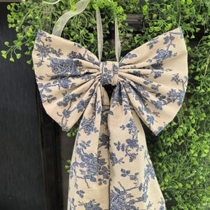 May include: A large decorative bow made of beige fabric with a blue floral pattern. The bow is hanging from a white ribbon and is likely used for home decor or seasonal decoration. The bow is centered in the frame.