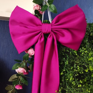 May include: A large, bright fuchsia satin bow with long tails. The bow is hanging from a wreath of green foliage and pink flowers.