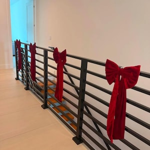 May include: A series of four deep red velvet bows attached to a black metal horizontal railing. Each bow features a large, multi-looped knot with two long, hanging fabric tails that drape vertically down the front of the railing structure.