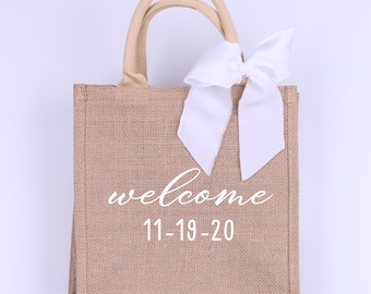 May include: A brown burlap tote bag with white handles and a white bow. The bag has the text "welcome 11-19-20" printed on the front.