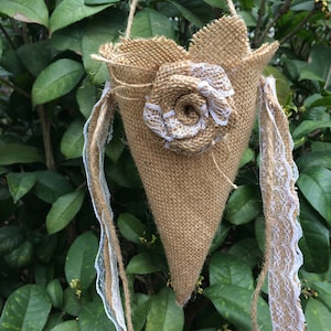 Puede incluir: Una bolsa de yute en forma de cono con una flor de encaje blanco y una cinta de encaje blanco que cuelga de ella. La bolsa está colgada de una rama de una planta verde frondosa.