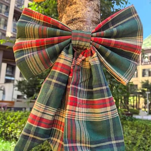 May include: A large, decorative plaid bow in shades of green, red, and gold is tied around a tree trunk. The bow's fabric features a classic tartan pattern, with long, flowing tails. The bow is a festive decoration.
