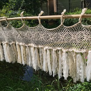 以下が含まれることがあります： 複雑な透かし編み模様と花柄が特徴のクリーム色のクロシェ編みバランス。下端には長い布のストリップとレースのリボンが連なって垂れ下がっており、水平な木製のロッドにループで吊り下げられています。