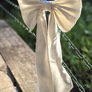 May include: A large, off-white fabric bow tied around a green metal post. The bow has long, flowing tails and is set against a blurred background of a wooden surface, a chain, and greenery. The bow is made of a textured fabric.
