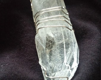 Puede incluir: Un colgante de cristal de cuarzo transparente envuelto en alambre plateado. El cristal es alargado con una textura natural rugosa. El alambre está en bucle en la parte superior para colgarlo. El colgante se muestra sobre un fondo oscuro.