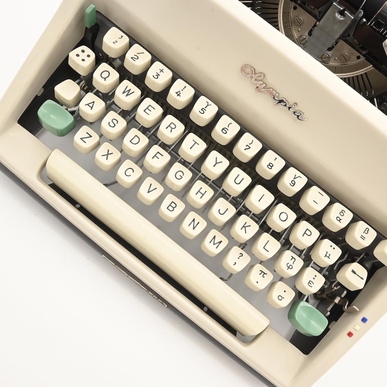 May include: An off-white Olympia typewriter featuring a QWERTY keyboard layout with cream-colored keys, black lettering, and two light green modifier keys. The metal frame includes a silver Olympia logo and a long space bar at the bottom.