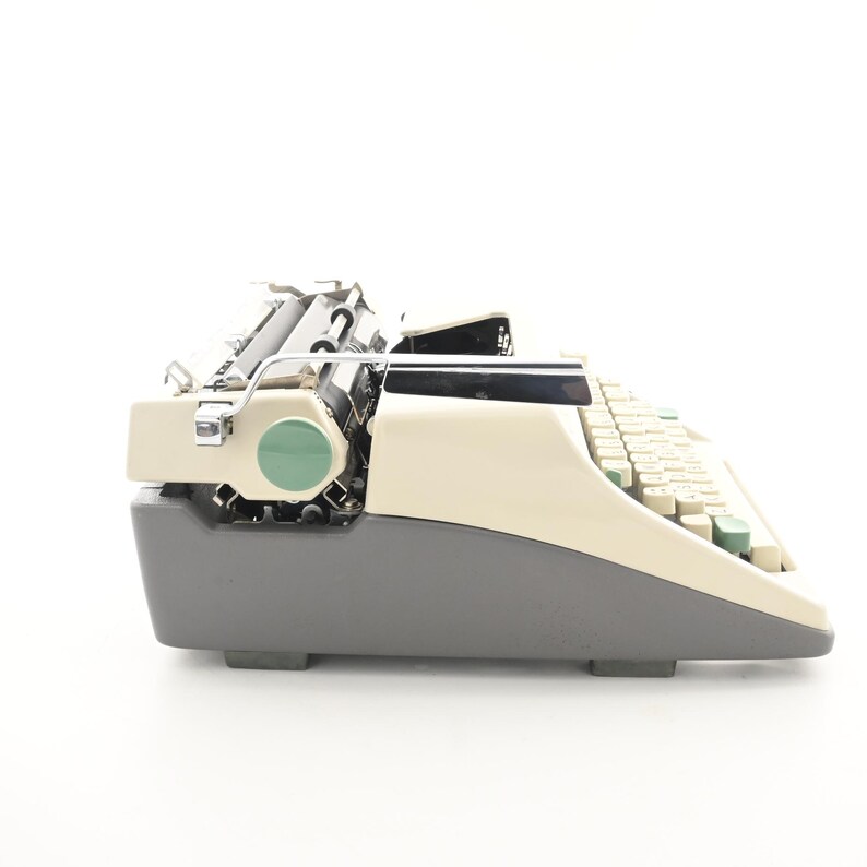 May include: A vintage manual typewriter featuring a two-tone design with a cream-colored top casing and a dark gray base. The machine includes a black carriage, a green circular accent on the side, and a keyboard with light-colored keys.