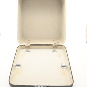 May include: An open, empty, square-shaped hard-shell carrying case with a dark gray exterior and a light beige interior. The case features metal hinges connecting the lid to the base, along with four small metal mounting brackets inside the base.