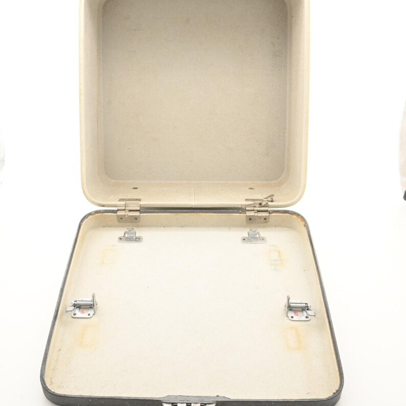 May include: An open, empty, square-shaped hard-shell carrying case with a dark gray exterior and a light beige interior. The case features metal hinges connecting the lid to the base, along with four small metal mounting brackets inside the base.