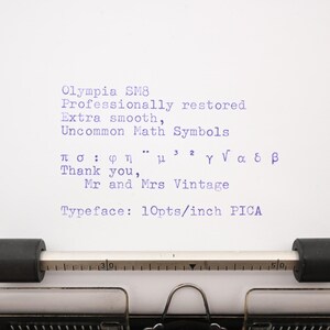May include: A white sheet of paper in a typewriter with blue ink text reading Olympia SM8, Professionally restored, Extra smooth, Uncommon Math Symbols, various Greek and math characters, Thank you, Mr and Mrs Vintage, Typeface: 10pts/inch PICA.