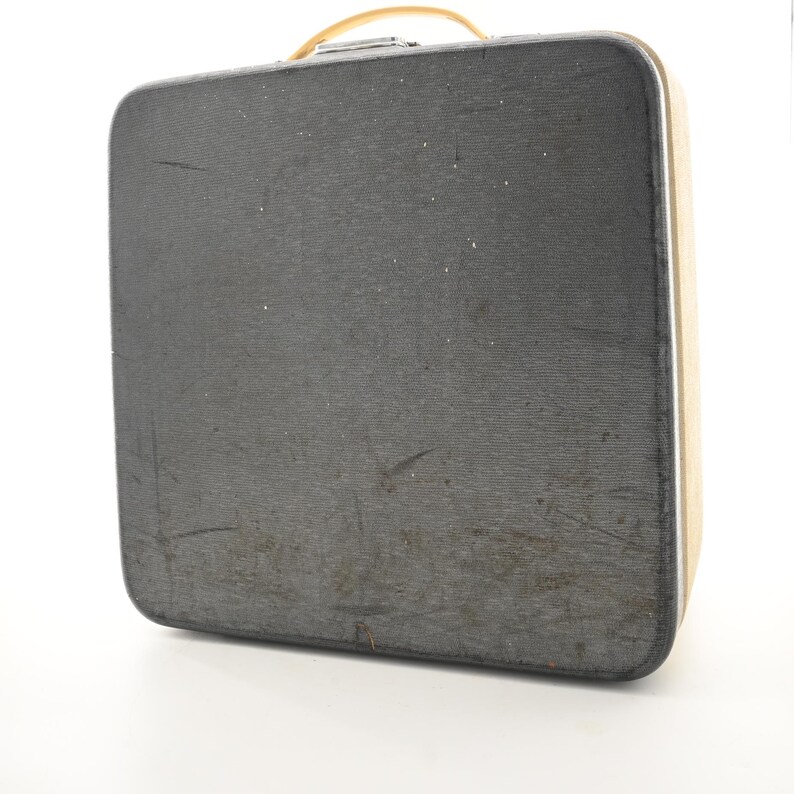 May include: A rectangular hard-shell suitcase with a dark gray textured exterior and a light tan side panel. The case features a light tan top handle and a silver-toned metal latch mechanism. The surface shows visible signs of wear and light scratches.