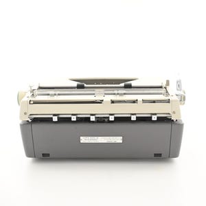 May include: A vintage typewriter featuring a two-tone design with a cream-colored top section and a dark gray base. A rectangular metal identification plate is centered on the front, displaying the text OLYMPIA WERKE AG WILHELMSHAVEN.