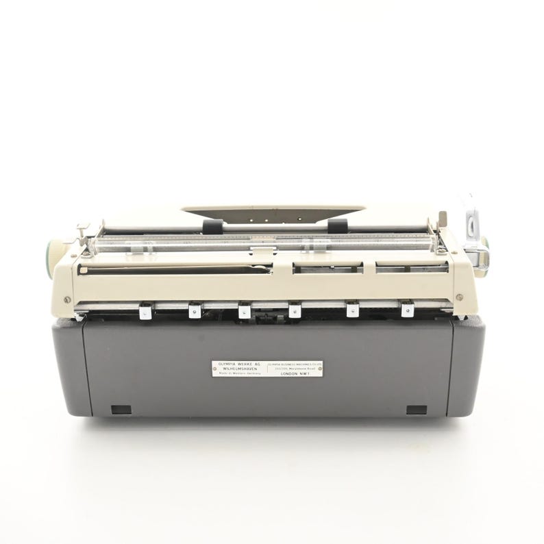 May include: A vintage typewriter featuring a two-tone design with a cream-colored top section and a dark gray base. A rectangular metal identification plate is centered on the front, displaying the text OLYMPIA WERKE AG WILHELMSHAVEN.