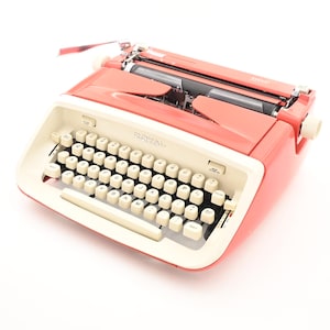 May include: A vintage coral Royal typewriter with a cream keyboard. The typewriter has a standard QWERTY layout with black keys and white lettering. The Royal logo is visible on the front.
