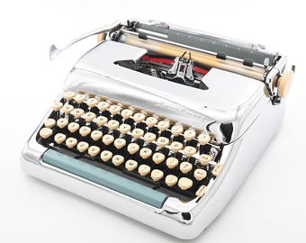 May include: A vintage Smith-Corona typewriter with a shiny chrome finish. The typewriter has a full keyboard with off-white keys, a blue front panel, and a red ribbon visible inside. The carriage return lever is on the left side.