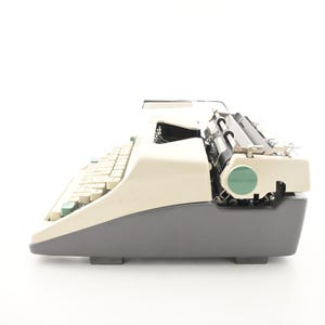 May include: A side view of a vintage typewriter featuring a two-tone design with a cream-colored top casing and a dark gray base. The machine includes a series of cream keys, a black platen roller, and a circular green accent on the side panel.