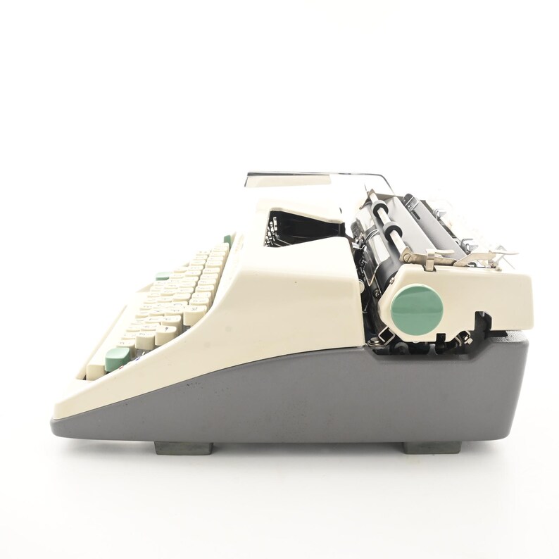 May include: A side view of a vintage typewriter featuring a two-tone design with a cream-colored top casing and a dark gray base. The machine includes a series of cream keys, a black platen roller, and a circular green accent on the side panel.