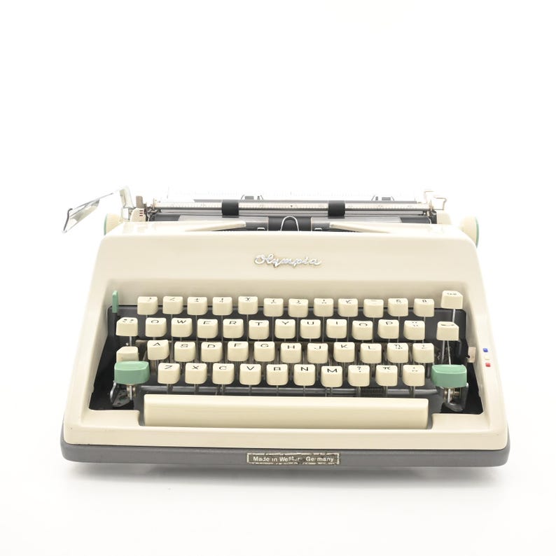 May include: An off-white Olympia typewriter with a dark gray base and light green accent keys. The keyboard features white keys with black lettering, a wide space bar, and a metal carriage. A small label at the bottom reads Made in West Germany.