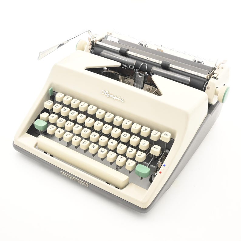 May include: An off-white Olympia typewriter with a grey base and light green accent keys. The QWERTY keyboard features cream-colored circular keys with black lettering, a metal carriage, and a paper bail. The brand name Olympia is printed in script.