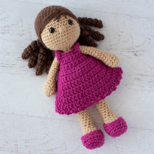 May include: A crocheted doll with brown hair wearing a pink dress. The doll has brown eyes and a beige face.