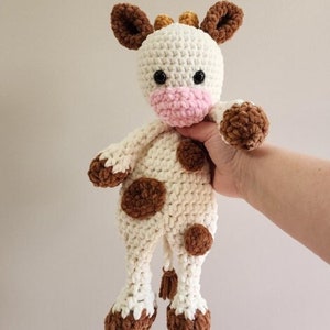 May include: A crocheted stuffed cow toy with brown spots and a pink nose. The cow is white with brown accents and has a long tail with a tassel.