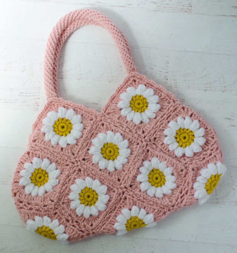 May include: A pink crocheted tote bag with white and yellow daisy flowers. The bag has two straps and is made with a granny square stitch.