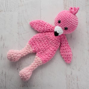 May include: A handmade pink crochet flamingo toy with a white and black beak, black eyes, and a small tuft of feathers on its head. The flamingo's legs and feet are a lighter shade of pink. The toy is lying on a white wooden surface.