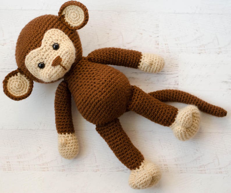 May include: A brown crocheted monkey toy with beige ears, hands, and feet. The monkey is lying on its back with its legs and arms outstretched.