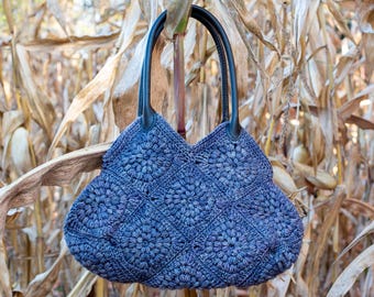 May include: A blue crocheted tote bag with black leather handles. The bag is hanging from a stalk of dried corn.
