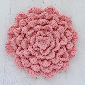 May include: A pink crocheted flower with a textured, layered design. The flower is made with a single strand of yarn and has a circular shape.