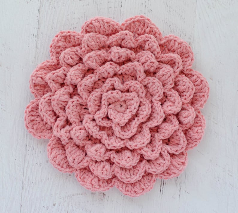 May include: A pink crocheted flower with a textured, layered design. The flower is made with a single strand of yarn and has a circular shape.