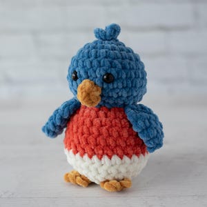 以下が含まれることがあります： 青、赤、白、茶色の手編みかぎ針編み鳥。鳥は青い頭と翼、赤い胸、白いお腹、茶色のくちばしと足を持っています。柔らかく、質感のある糸で作られており、黒い安全な目があります。