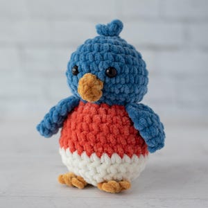 May include: A handmade, plush bird toy with a blue head and wings, a red chest, and a white belly. The bird has a small, orange beak and feet, and black eyes. The toy is made of a soft, textured material.