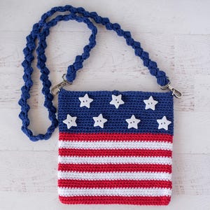 May include: A crocheted crossbody bag with a blue strap and a design resembling the American flag. The bag features a blue top with white star-shaped embellishments and red and white stripes below. The strap is a twisted blue cord.