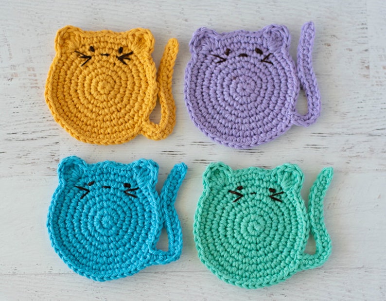 May include: Four crocheted cat-shaped coasters in yellow, purple, blue, and green. Each coaster has a tail and a face with embroidered whiskers.