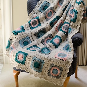 May include: A crocheted throw blanket with a granny square pattern in shades of gray, teal, and pink. The blanket is draped over a wooden armchair.