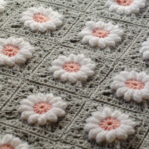 May include: A crocheted blanket with a repeating pattern of white daisies with pink centers on a gray background.