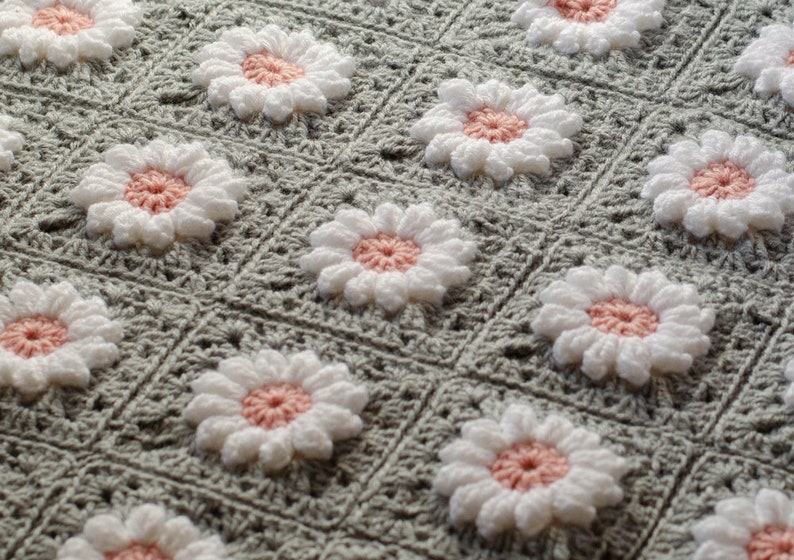 May include: A crocheted blanket with a repeating pattern of white daisies with pink centers on a gray background.