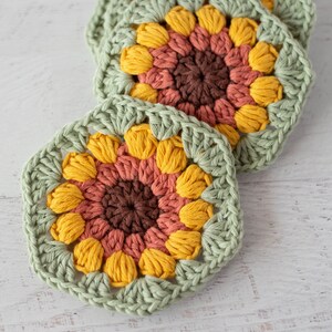 Op de afbeelding: Drie gehaakte zonnebloemonderzetters in tinten groen, geel, oranje en bruin. De onderzetters zijn gemaakt met een granny square-patroon.