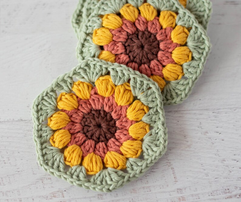Op de afbeelding: Drie gehaakte zonnebloemonderzetters in tinten groen, geel, oranje en bruin. De onderzetters zijn gemaakt met een granny square-patroon.