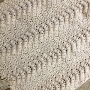 May include: A crocheted throw blanket with a chevron pattern in a cream color. The blanket has a fringe border.