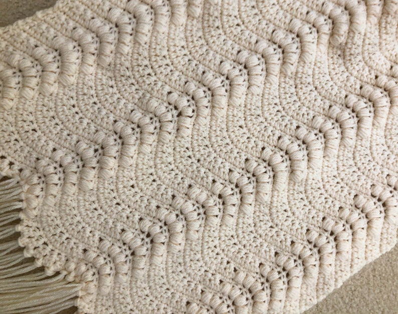 May include: A crocheted throw blanket with a chevron pattern in a cream color. The blanket has a fringe border.