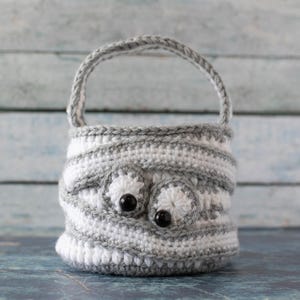May include: A gray and white crocheted basket with a handle and two black eyes. The basket is made with a striped pattern.