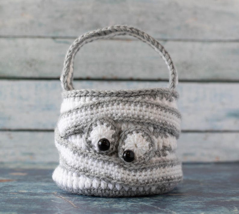 May include: A gray and white crocheted basket with a handle and two black eyes. The basket is made with a striped pattern.